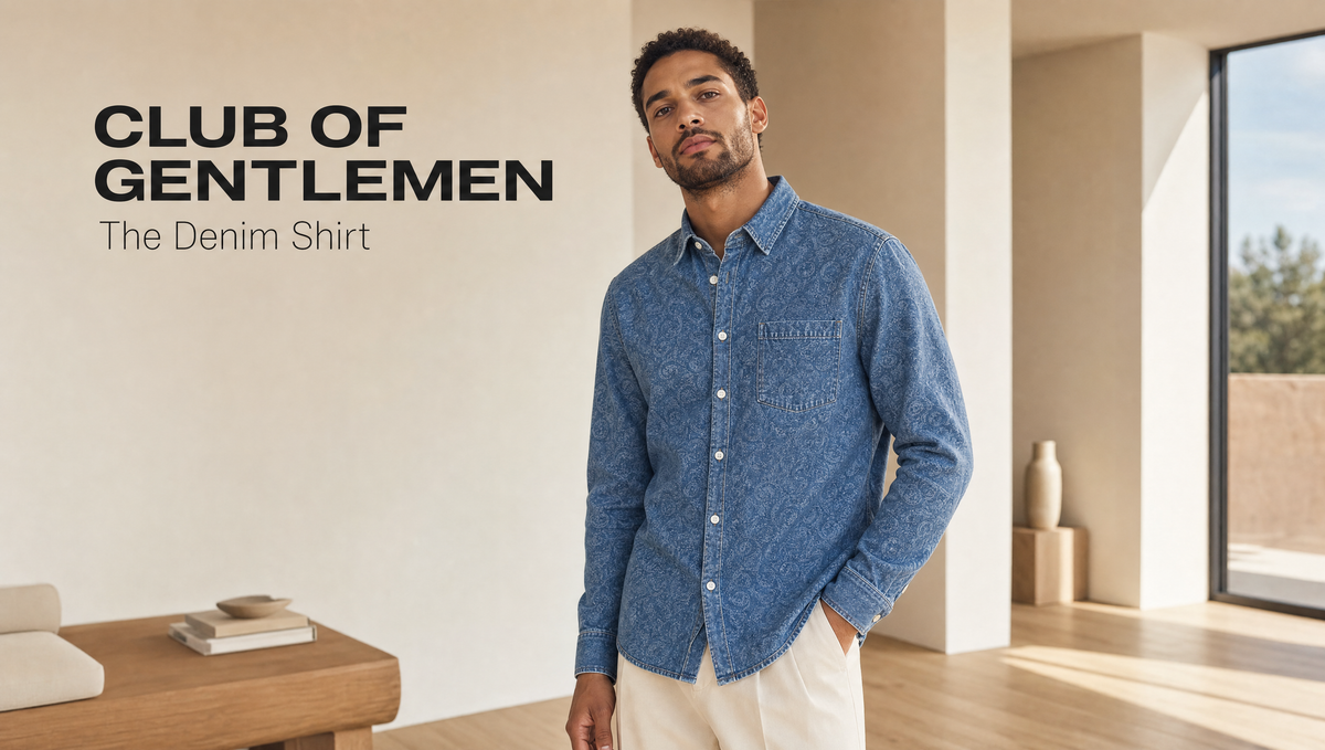 A man wearing a blue patterned long-sleeve denim shirt and cream pants stands in a modern interior setting. Text on the left reads 'CLUB OF GENTLEMEN The Denim Shirt'.