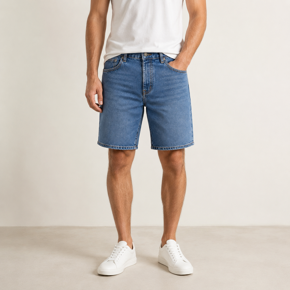 A man wearing medium-wash blue denim shorts, a white t-shirt, and white sneakers, standing against a plain off-white studio background.