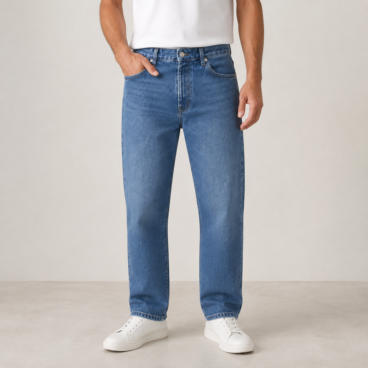 A person wearing medium-wash blue jeans with a white t-shirt and white sneakers, standing against a neutral light beige studio background.